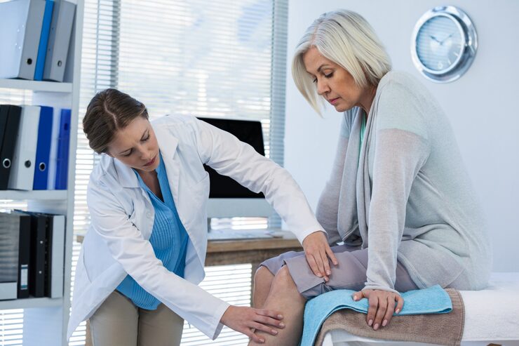 A physiotherapist (or clinician) in a white coat leans over to gently examine the knee of an older female patient who is sitting on an examination table. The physiotherapist's hands are placed on the woman's knee and lower leg, indicating a detailed joint assessment. This visualizes the crucial stage of the WSIB physiotherapy assessment report for a knee injury, which is vital documentation for claim approval.