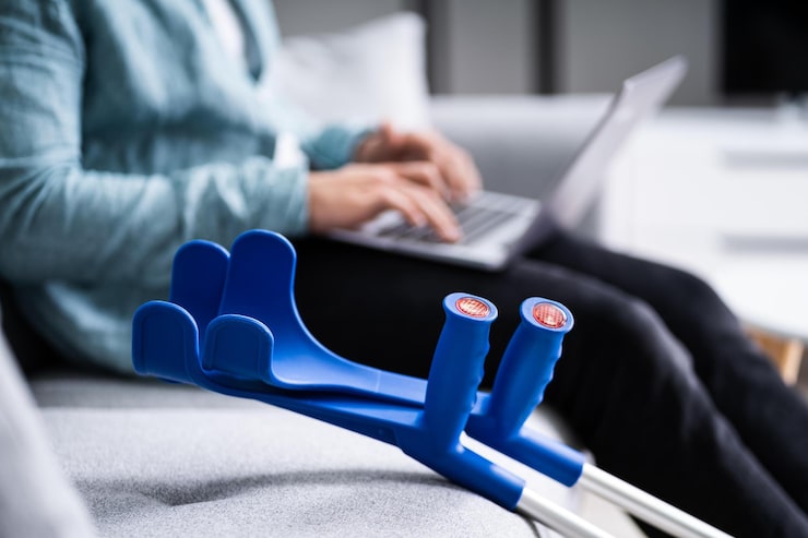 A shallow-focus image showing a pair of bright blue crutches resting on a grey couch in the foreground. In the soft-focus background, a person with an injury, wearing a light blue shirt, is seen sitting and typing on a laptop. This represents the process of remotely managing or following up on a WSIB claim (such as filing WSIB Form 6 or checking WSIB claim status) while using mobility aids due to a work-related injury.