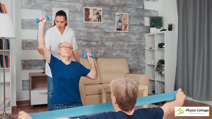 A female physiotherapist helps two seniors in a living room doing at-home exercises.