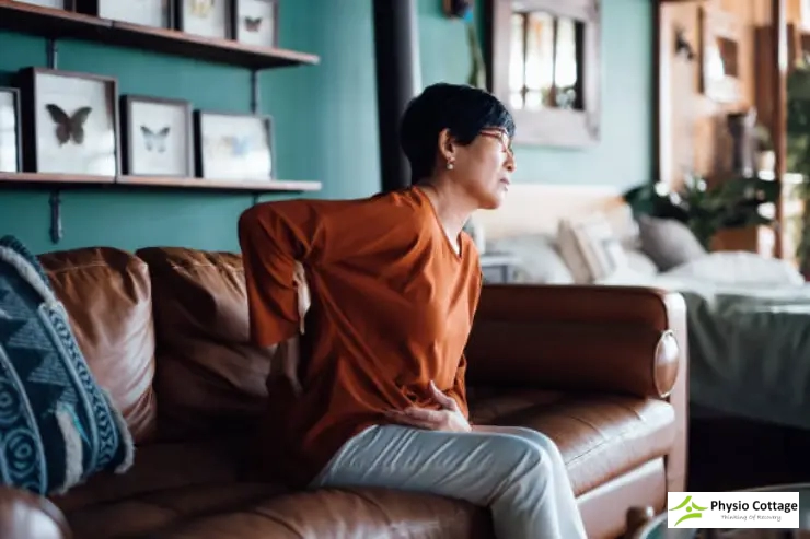Woman sitting on a couch gripping her lower back, illustrating acute sciatica pain.