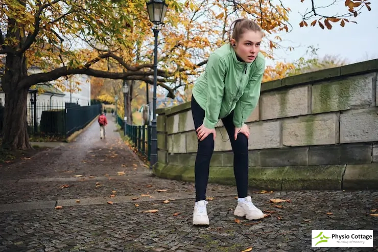 Woman in a green jacket pausing and resting her hands on her knees after a walk.