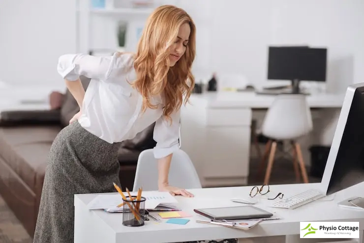 Woman in office standing and stretching, grimacing from lower back pain.