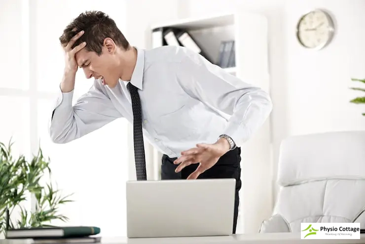 Man standing at desk in pain, bending over a laptop and holding his head.